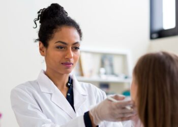 A clinician wearing a white coat and gloves examines a patient’s face during a consultation.